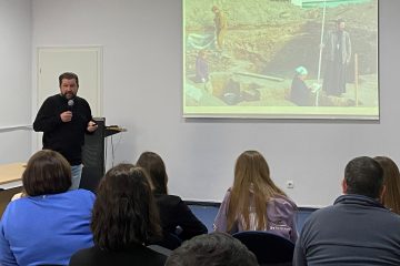 Лектор с микрофоном выступает перед аудиторией, на экране – фото археологических работ.