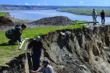 Археологи работают в раскопе на краю обрыва над Иртышем, видны деревянные постройки села.