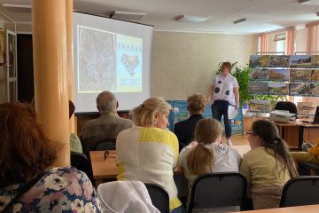 Лектор у экрана с иллюстрациями, на переднем плане сидят слушатели разных возрастов.